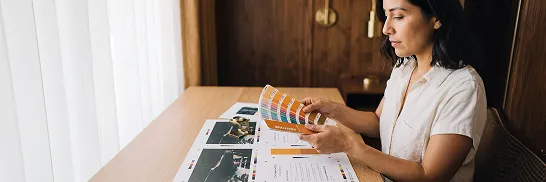 Graphic designer carefully reviewing freshly printed proof sheets alongside a Pantone color fan at a design desk with warm morning window light Graphic designer carefully reviewing freshly printed proof sheets alongside a Pantone color fan at a design desk with warm morning window light