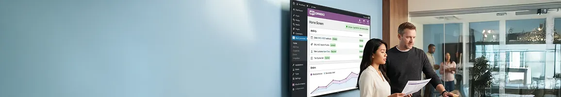 Two team members reviewing WooCommerce integration sync metrics on large screens, side angle, natural workplace light Two team members reviewing WooCommerce integration sync metrics on large screens, side angle, natural workplace light